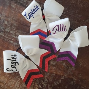 May include: Three white cheer bows with glitter accents. The bows have different names written on them: "Captain", "Alli", and "Eagles".