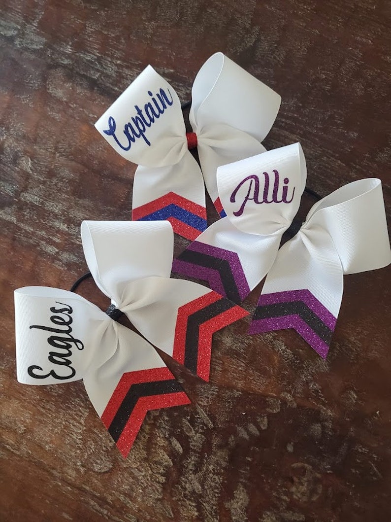 May include: Three white cheer bows with glitter accents. The bows have different names written on them: "Captain", "Alli", and "Eagles".