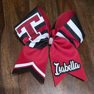 May include: A red, black, and white cheerleading bow with the name "Isabella" in white script. The bow features a glittery "T" and a rhinestone center. The bow is made of ribbon.