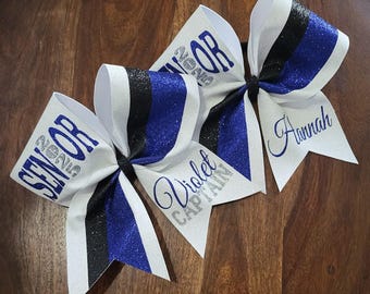Może przedstawiać: Trzy białe kokardy do cheerleadingu z brokatowymi akcentami. Każda kokarda ma czarno-niebieski pasek, z napisami "SENIOR 2023", "Violet CAPTAIN" i "Hannah". Kokardy są ułożone na drewnianej powierzchni.