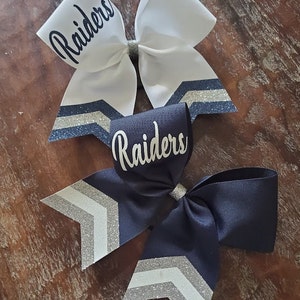 May include: Two cheer bows with a chevron pattern and silver glitter accents. The bows are white and navy blue with the word "Raiders" written on each bow.