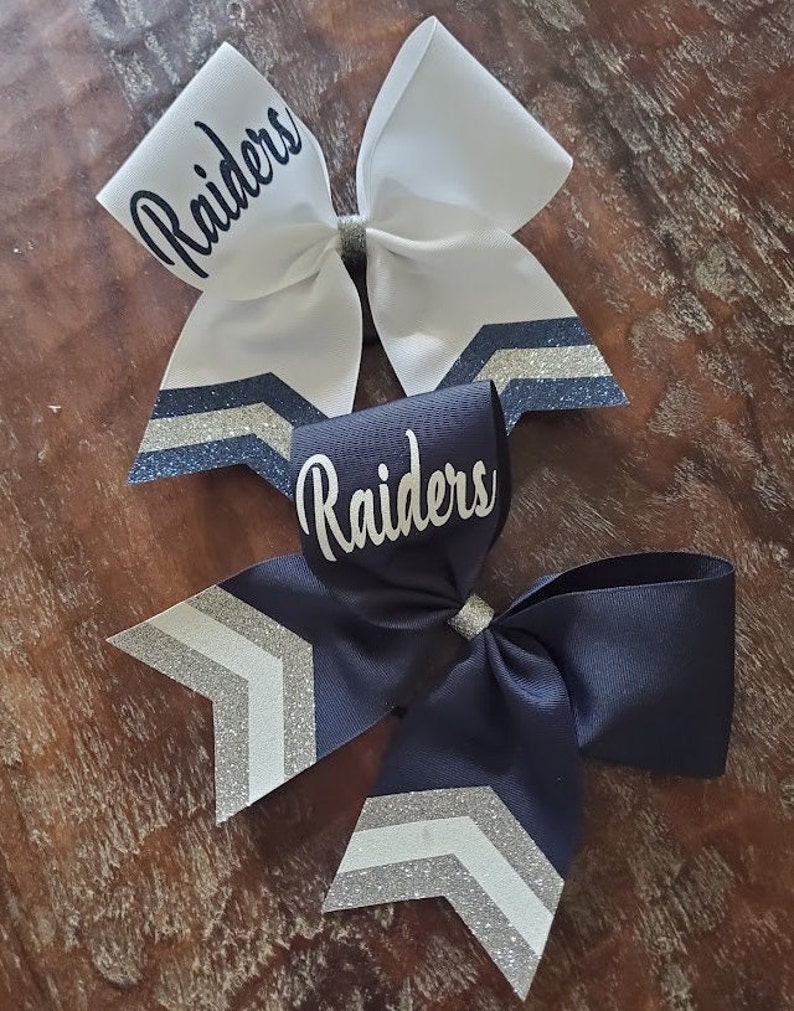 May include: Two cheer bows with a chevron pattern and silver glitter accents. The bows are white and navy blue with the word "Raiders" written on each bow.