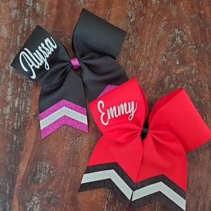 May include: Two personalized cheer bows with black and white chevron accents. The top bow is black with a pink chevron and the name "Alyssa" in white. The bottom bow is red with a white chevron and the name "Emmy" in white.