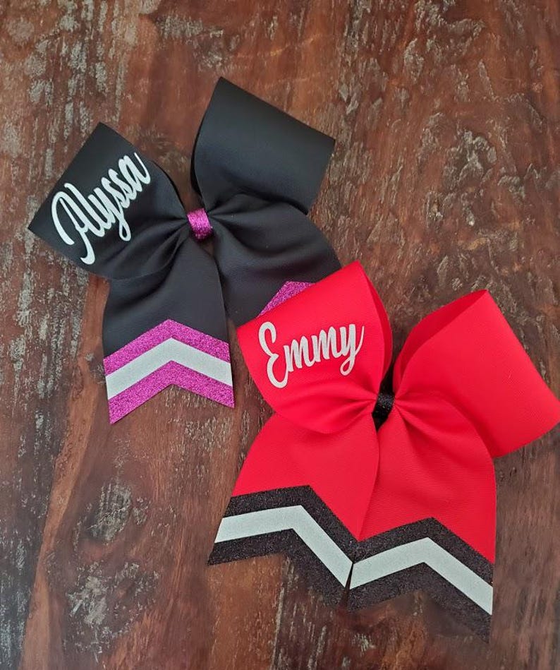 May include: Two personalized cheer bows with black and white chevron accents. The top bow is black with a pink chevron and the name "Alyssa" in white. The bottom bow is red with a white chevron and the name "Emmy" in white.