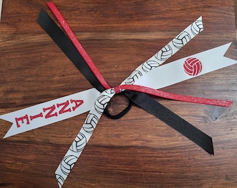 May include: White and red ribbon hair tie with black ribbon and a volleyball design. The ribbon has the text "E-IZZA" printed on it.