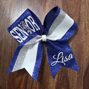 May include: A large hair bow featuring royal blue glitter fabric, a silver glitter stripe, and a white stripe. The left side displays the word SENIOR in white block letters, and the right side features the name Lisa in white script.