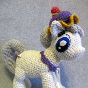 May include: White crocheted pony with purple and yellow accents. The pony has a purple hat with a pink pom-pom on top. The pony is wearing a purple and white ribbon around its neck.