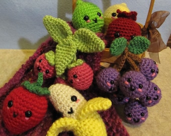 May include: A selection of crocheted fruit toys, including cherries, strawberries, a banana, grapes, a lemon, and a lime, displayed in a small wooden basket and on a purple surface. Each fruit has a smiling face and is made with colourful yarn.