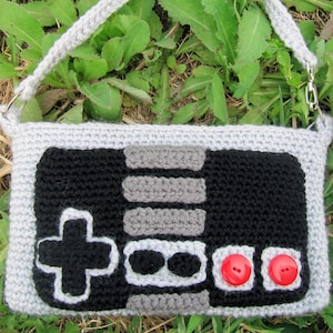 May include: A gray crocheted purse shaped like a vintage video game controller. The controller has black buttons and a gray and black directional pad.