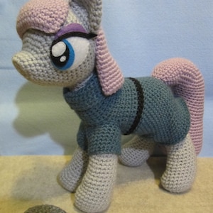 May include: A gray crocheted pony with pink and purple accents, wearing a blue sweater with a black stripe. The pony is standing on a beige surface with a gray rock in front of it.