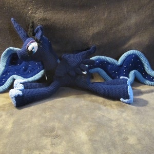May include: A crocheted plush toy of a blue and black unicorn with a blue and white mane and tail. The unicorn is lying on its side with its head turned to the side.