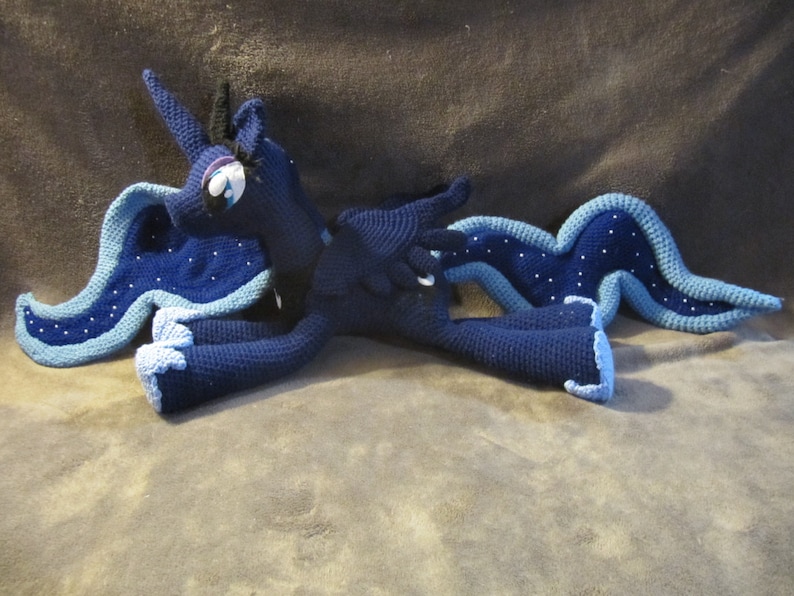 May include: A crocheted plush toy of a blue and black unicorn with a blue and white mane and tail. The unicorn is lying on its side with its head turned to the side.