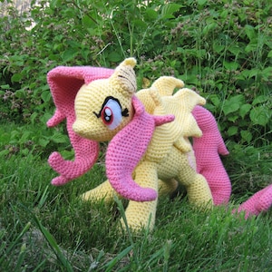 May include: A crocheted plush toy of a yellow and pink pony with a pink mane and tail. The pony is standing on green grass.