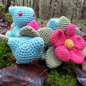 Può includere: Due giocattoli di peluche all'uncinetto a forma di fiore e una creatura blu con occhi rossi. I giocattoli sono seduti su un letto di muschio e foglie.