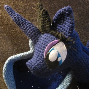 May include: A crocheted plush toy of a blue unicorn with a black mane and tail. The unicorn has a purple and white eye with a blue tear.