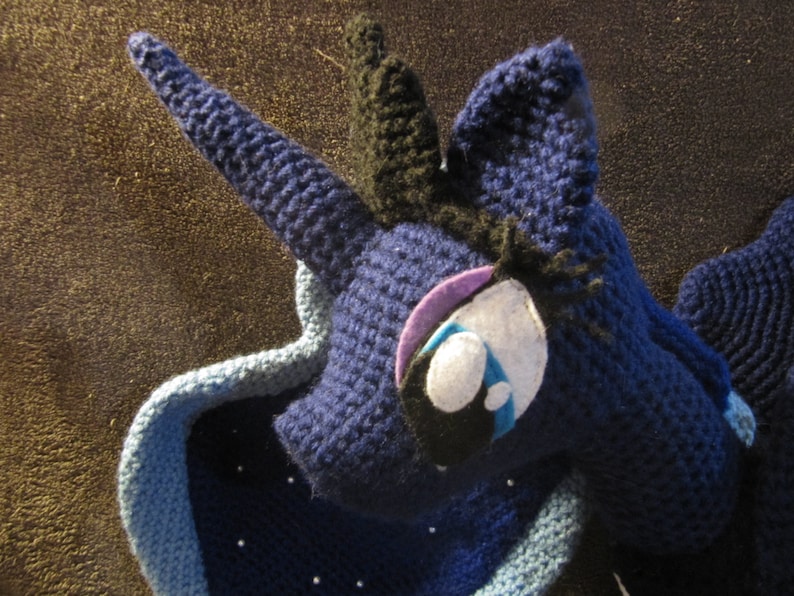 May include: A crocheted plush toy of a blue unicorn with a black mane and tail. The unicorn has a purple and white eye with a blue tear.