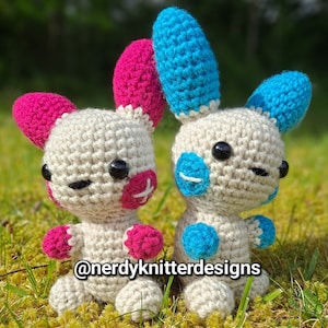 May include: Two crocheted amigurumi toys with white bodies, pink and blue accents, and black eyes. The toy on the left has a pink pacifier and pink arms and legs. The toy on the right has blue arms and legs. The toys are sitting on a bed of green moss. The text "@nerdyknitterdesigns" is visible below the toys.
