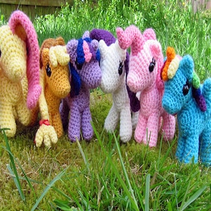 Puede incluir: Siete figuritas de pony de crochet en varios colores, incluyendo amarillo, marrón, morado, blanco, rosa, azul y arcoíris. Los ponis están de pie sobre un lecho de hierba verde.