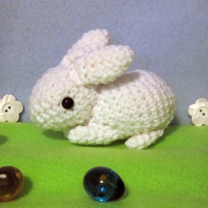 Könnte beinhalten: Ein weißes Häkel-Hasenspielzeug mit schwarzen Augen. Der Hase sitzt auf einer grünen Oberfläche mit mehreren kleinen, bunten Glasmurmeln und weißen, blumenförmigen Knöpfen.