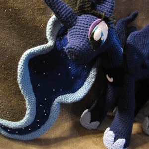 May include: A blue and black crocheted plush toy of a unicorn with a long, flowing mane and tail. The unicorn has a white star on its forehead and a blue eye.