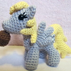 May include: A crocheted gray and yellow pony toy with a brown nose and a single black eye. The pony is holding a brown chocolate bar in its mouth.