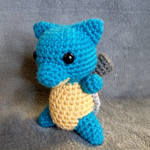 May include: A crocheted blue and yellow Squirtle plush toy with black eyes. The toy is standing on a gray fuzzy surface.