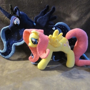 May include: Three crocheted My Little Pony figures. The pony on the left is blue and purple with a star pattern. The pony in the middle is yellow with a pink mane and tail. The pony on the right is pink with a yellow mane and tail.