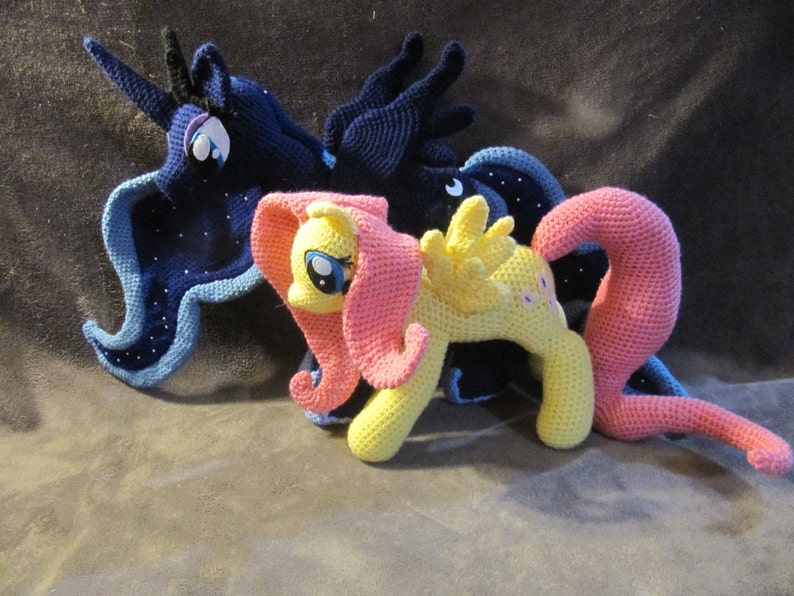 May include: Three crocheted My Little Pony figures. The pony on the left is blue and purple with a star pattern. The pony in the middle is yellow with a pink mane and tail. The pony on the right is pink with a yellow mane and tail.