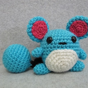 May include: A blue and white crocheted plush toy with red ears. The toy is shaped like a small animal with a round body and a long tail.