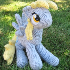 May include: A crocheted plush toy of a light blue pony with yellow hair and wings. The pony has large, black eyes with yellow pupils and is sitting on a green grassy surface.