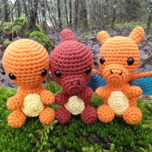 Könnte beinhalten: Drei gehäkelte Plüschtiere in Form eines feuerspeienden Drachen. Der Drache links ist orange mit gelbem Bauch, der mittlere Drache ist braun mit gelbem Bauch und der Drache rechts ist orange mit gelbem Bauch und blauen Flügeln.