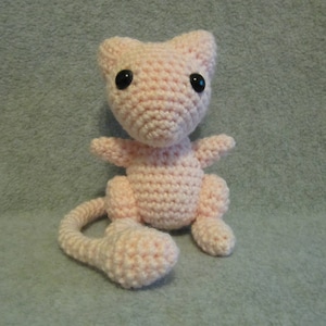 May include: A pink crocheted plush toy resembling a Pokemon character with black eyes. The toy has a long tail and is sitting on a gray surface.