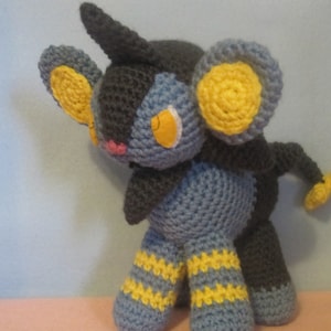 May include: A crocheted plush toy of a blue and black Pokémon with yellow accents. The toy has a long, pointed tail and large, yellow ears.
