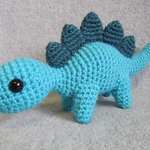 Op de afbeelding: Een gehaakt blauw dinosaurus speelgoed met een stekelig rug in teal groen. De dinosaurus heeft zwarte ogen en een kleine, ronde snuit.