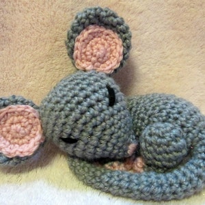May include: A gray crocheted mouse with pink ears and a curled tail, sleeping on a white fuzzy surface.