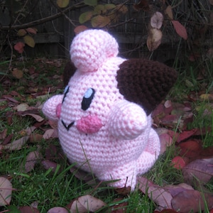 May include: A pink and brown crocheted plush toy resembling a Pokemon character. The toy has a round body, large eyes, and a small brown ear.