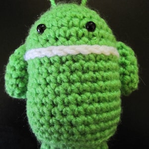 May include: Green crocheted plush toy shaped like the Android mascot. The toy has black eyes and a white crocheted mouth.