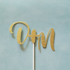 May include: A gold glitter cake topper with the letters "DH M" in a cursive font. The letters are attached to a white stick, set against a light blue background. This decorative item is suitable for cakes and other baked goods.