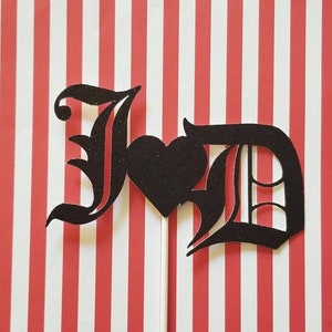 May include: Black cake topper with the letters "JD" in a gothic font with a heart between the letters.