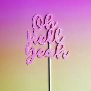 May include: Pink glitter cake topper with the text "oh hello yeah" on a white stick.