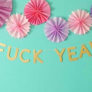 Może przedstawiać: Złota błyszcząca girlanda z napisem "F**K YEAH" wisząca na turkusowym tle z różowymi i fioletowymi papierowymi ozdobami.
