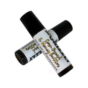 Può includere: Due flaconi roll-on in vetro ambrato con tappi neri, posizionati a forma di X. Ogni flacone presenta un'etichetta bianca con il testo Grizzly, Corn Bread & Honey Butter e Pick-Me-Up Refresher stampato in caratteri neri e dorati.