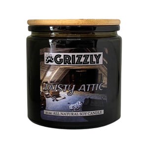 Puede incluir: Un tarro de vela de vidrio negro con tapa de madera. El tarro tiene una etiqueta que dice "Grizzly" y "Dusty Attic" con una imagen de un ático. La etiqueta también dice "11 oz. All Natural Soy Candle".