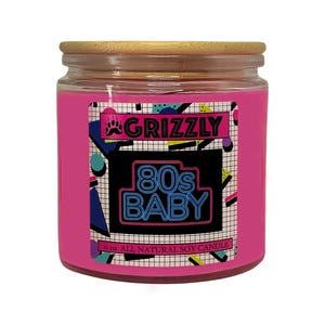 May include: Pink soy candle in a glass jar with a wooden lid. The label features a black and white geometric design with the text "Grizzly 80's Baby" in neon blue and pink. The candle is 11 oz and is all natural soy.