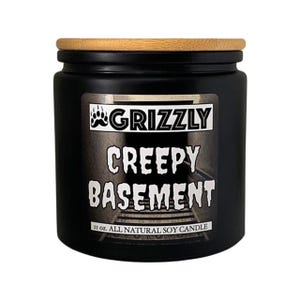 Puede incluir: Vela de soja negra con tapa de madera. La etiqueta dice "Grizzly Creepy Basement" y "11 oz. All Natural Soy Candle".