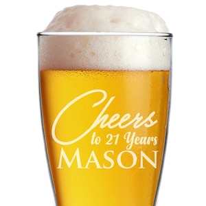May include: A tall, clear glass beer stein with a white inscription that reads "Cheers to 21 Years MASON". The glass is filled with a golden-colored beer and topped with a white head of foam.