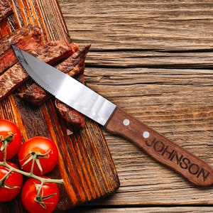 May include: A wooden cutting board with a steak sliced into thin pieces, a knife with a wooden handle engraved with the word "JOHNSON", and red tomatoes on a vine.