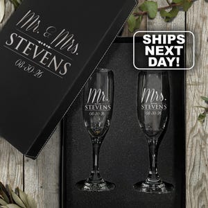 May include: A black gift box containing two clear glass champagne flutes. The box lid and the glasses are engraved with the text Mr. & Mrs. STEVENS and the date 08-30-26. The glasses are set into a black foam insert within the rectangular box.