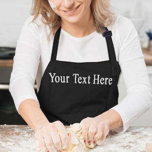 May include: Black apron with the text "Your Text Here" in white. The apron is being worn by a person in a kitchen, kneading dough. The person is wearing a white long-sleeved shirt.