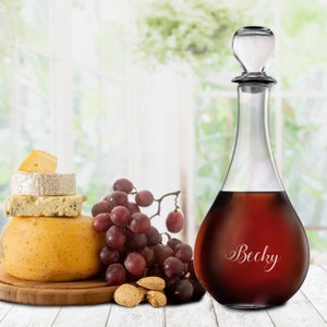 May include: A clear glass decanter with a red liquid inside. The decanter has a stopper and is engraved with the name "Becky". The decanter is sitting on a wooden surface with grapes, cheese, and almonds.
